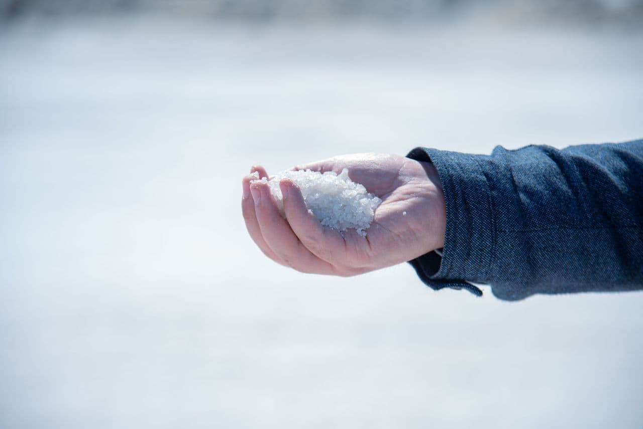 A hand holding white de-icer granules over an icy surface