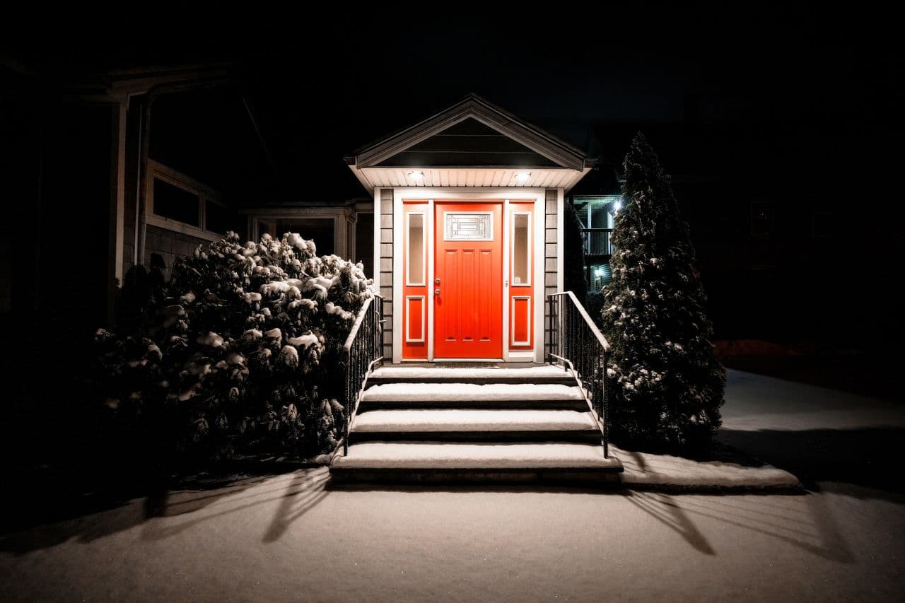 Snow lying on the front steps of a lit porch with a red door on a winter night