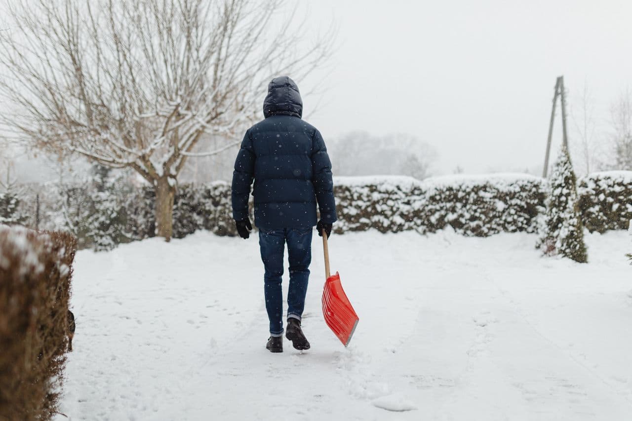 A person in a puffer jacket carrying a snow shovel on a winter day